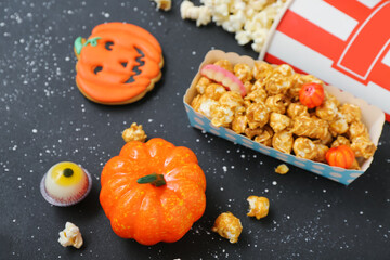 Paper box with tasty caramel popcorn, cookie and pumpkin on dark background, closeup. Halloween celebration