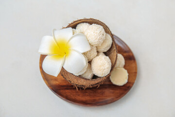 Fototapeta na wymiar Coconut shell with white chocolate candies and plumeria flower on light table