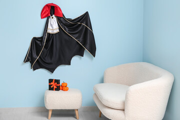 Sofa, pouf with gift box for Halloween, pumpkins and cloak hanging on blue wall in living room