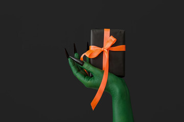 Green hand of witch with gift box on dark background. Halloween celebration