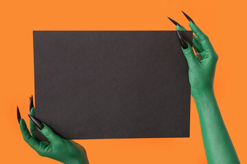 Hands of witch with blank black paper sheet on orange background. Halloween celebration