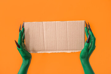 Green hands of witch with blank paper sheet on orange background. Halloween celebration
