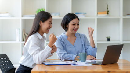 Two asian businesswoman expressing happiness in workplace smiling and raising hands in joy with laptop computer in modern office.