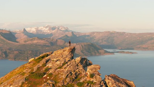 Male hiker on R&oslash;d&oslash;ya mountain top in arctic Norway midnight sun; drone orbit