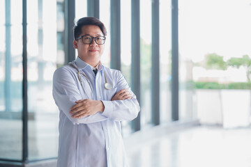 Horizontal waist-up portrait of a handsome male Arab Malay doctor, wearing white coat, standing with arms crossed outdoors on the background of clinic building. Close up, copy space