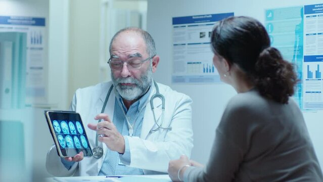 Senior male doctor showing MRI brain scan on digital tablet and explaining the diagnosis to female patient during medical appointment in clinic