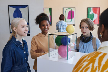 Diverse group of smiling teenagers listening to female teacher or tour guide in modern art gallery