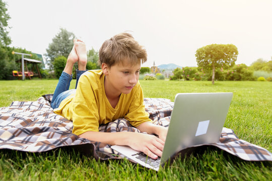TTeen boy with a laptop sits in a park. School boy is typing on computer outdoors. Young boy studying online with a computer. Boy is online lesson while outside.