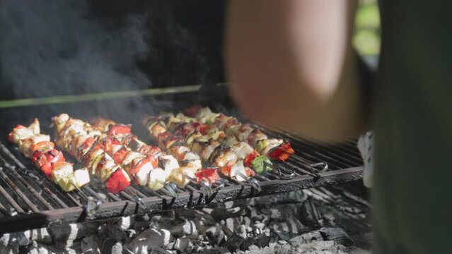 Cooking outdoors barbecue as cook flips chicken skewers on the hot grill. Smoking and hot brochettes are turned to other side to be roasted on coal grill in backyard BBQ party or camping site cookout