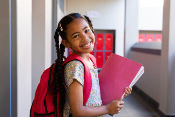 Portrait of happy biracial schoolgirl with books and school bag at elementary school corridor