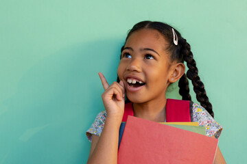 Happy biracial schoolgirl with books wearing school bag over blue background at elementary school