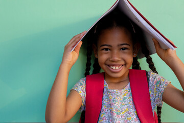 Portrait of happy biracial schoolgirl with book on head over blue background at elementary school