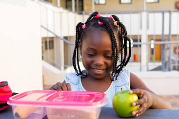 African american schoolgirl at table and having healthy lunch eating apple outside elementary school