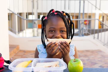 Fotobehang Happy african american schoolgirl having healthy lunch eating sandwich at elementary school © wavebreak3