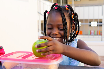 African american schoolgirl at table and having healthy lunch with apple outside elementary school
