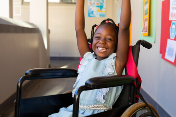 Portrait of african american schoolgirl sitting in wheelchair and raising hands at elementary school