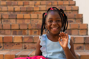Happy african american schoolgirl sitting on stairs waving hand at elementary school, copy space