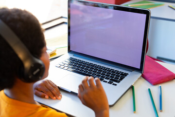 Focused biracial schoolboy with headphones using laptop with copy space in classroom