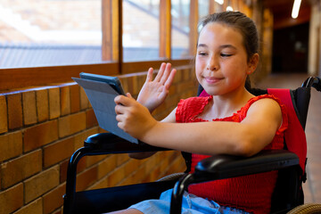 Happy caucasian schoolgirl sitting in wheelchair and using tablet at school corridor