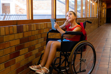 Caucasian schoolgirl sitting in wheelchair and using tablet at school corridor, copy space