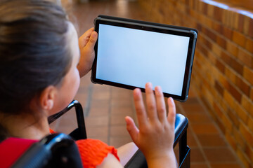 Caucasian schoolgirl sitting in wheelchair and using tablet with copy space at school corridor