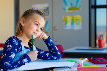Caucasian schoolgirl sitting at desk and thinking in classroom at elementary school, copy space