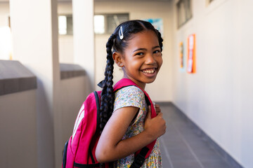 Portrait of happy biracial schoolgirl with school bag smiling in corridor at elementary school