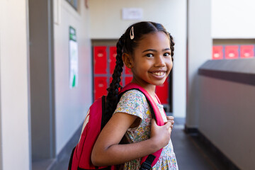 Portrait of happy biracial schoolgirl with school bag smiling in corridor at elementary school - Powered by Adobe