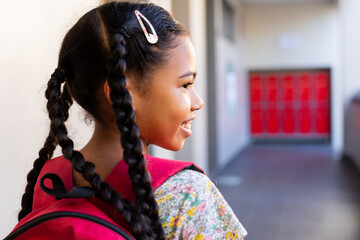 Happy biracial schoolgirl walking with school bag in corridor at elementary school