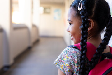 Happy biracial schoolgirl walking with school bag in corridor at elementary school, copy space