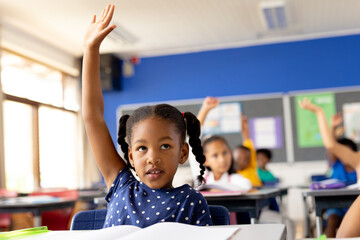 Diverse elementary schoolchildren sitting at desks and raising hands in class - Powered by Adobe