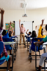 Diverse male teacher and elementary schoolchildren raising hands in class