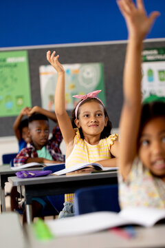 Diverse elementary schoolchildren sitting at desks and raising hands in elementary school class