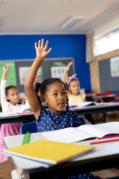 Diverse elementary schoolchildren sitting at desks and raising hands in class