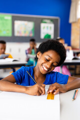 Happy african american elementary schoolboy writing in notebook at desk in class, copy space