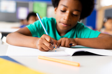 Portrait of happy african american elementary schoolboy writing in notebook at desk in class