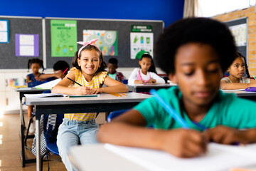 Portrait of biracial girl writing at desk with diverse elementary school class with copy space