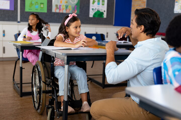 Happy biracial male teacher with girl in wheelchair in class at elementary school