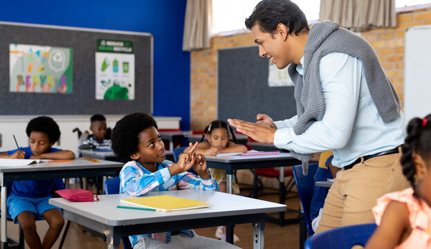 Diverse male teacher teaching school boy using sign language in class at elementary school