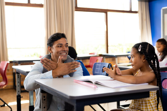 Diverse male teacher teaching school girl using sign language in class at elementary school