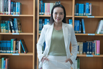 A female university lecturer in a business suit is searching for the book on the bookshelf in the...