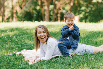Fototapeta premium Young Caucasian mother and her young son lie on a blanket on the grass in the park in summer and relax outdoors