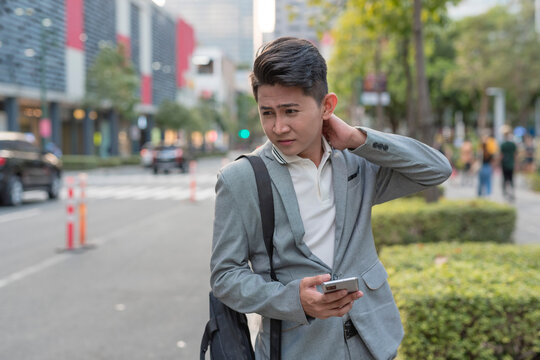 A young asian man suffers from a stiff neck from work while walking to a bus stop. Urban city setting.