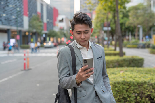 A young asian male office worker watches something on his phone while walking on the sidewalk after work.