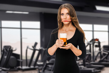 Pretty blonde girl posing with beer and sausage in modern gym while losing weight, concept against not healthy eating or healthy lifestyle with workouts