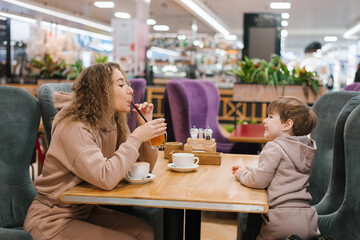Young curly haired mother and her little son are having breakfast or lunch in a cafe, sitting at a...