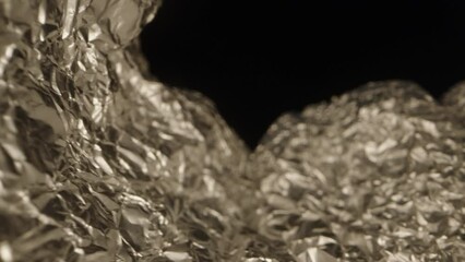 The camera moves along the creases of crumpled kitchen aluminum foil, highlighting its shiny texture against a black background. Macro shot.