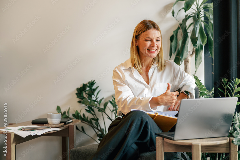 Wall mural smiling businesswoman talking with client via video call and showing thumb up sitting in coworking