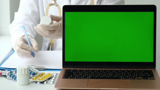 Laptop computer with green screen in medical clinic. Female doctor holding pills bottle and writing prescription on background. PC with blank and copy space chroma key monitor. Health care