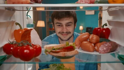 A young man looks into the refrigerator, examines its contents. A man with admiration looks at a plate with a sandwich. Choice between healthy and unhealthy food. View from inside the refrigerator.